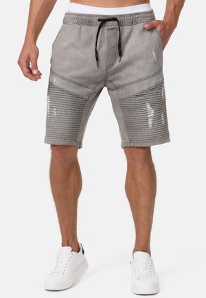 Homme portant un short gris côtelé et usé jusqu'aux genoux avec un cordon noir et des baskets blanches, debout les mains le long du corps.