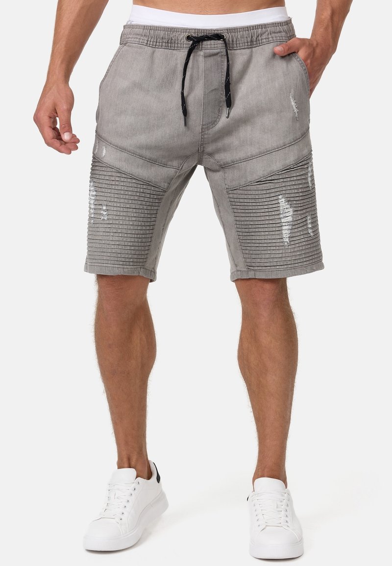Homme portant un short gris côtelé et usé jusqu'aux genoux avec un cordon noir et des baskets blanches, debout les mains le long du corps.