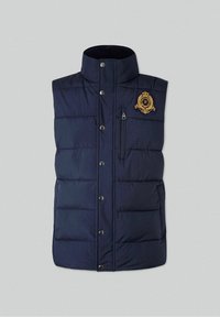 Veste matelassée bleu marine avec col haut, boutons-pression à l'avant, poche poitrine zippée et blason doré brodé sur le côté gauche de la poitrine.