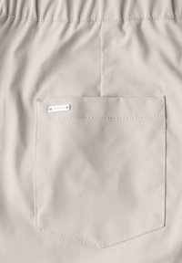 Ljusbeige tyg med en strukturerad yta, med en enda bakficka som har en rektangulär silveretikett fäst.