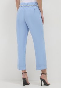 Donna che indossa pantaloni cropped blu chiaro con una cintura decorativa e sandali neri con tacco alto, in piedi su uno sfondo semplice.