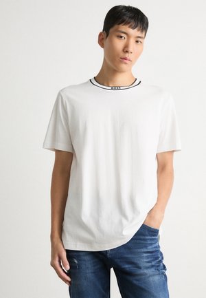 Camiseta de algodón blanca con mangas cortas y un cuello redondo con ribete negro. El logo "BOSS" se encuentra en el cuello. Se lleva con unos jeans azules.