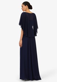 Frau mit geflochtenem Haar, das zu einem Dutt gebunden ist, trägt ein bodenlanges, marineblaues Kleid mit durchsichtiger Umhang-Überlage und steht im Profil vor einem weißen Hintergrund.