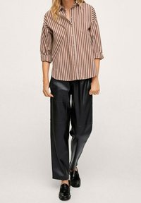 Chemise rayée marron avec un design à boutons, manches retroussées. Associée à un pantalon noir à jambes larges, brillant et des mocassins noirs.
