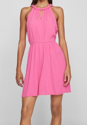 Kobieta w jaskrawo różowej sukience bez rękawów, z plecionym dekoltem halter i marszczoną talią, stojąca na szarym tle.