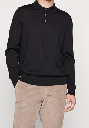 Homme portant un polo noir à manches longues et un pantalon beige en velours côtelé, debout avec une main dans la poche sur un fond uni.