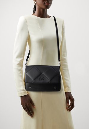 Sac à épaule en cuir noir matelassé avec logo, design plat, fermeture magnétique et sangle réglable. Modèle portant une longue robe crème.