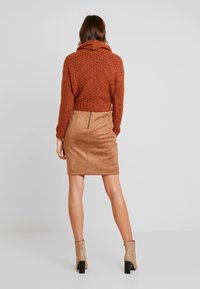 Un pull tricoté couleur rouille avec une capuche, associé à une jupe crayon en faux daim beige avec une fermeture éclair au dos. Des bottines marron complètent l'ensemble.