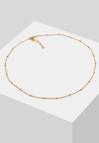 Collier délicat en chaîne dorée avec de petites perles rondes espacées régulièrement et un fermoir réglable sur une surface blanche et grise.