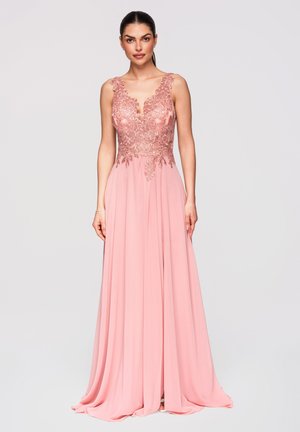 Femme debout portant une robe rose sans manches avec un corsage en dentelle fleurie et une jupe fluide, les cheveux tirés en une queue de cheval basse.