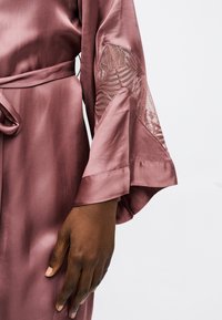 Σκούρο δέρμα χέρι που ακουμπά δίπλα στη μέση, με φόρεμα από σατέν σε αποχρώσεις μοβ με δαντελένιες εισαγωγές και μανίκια καμπάνα, πάνω σε ένα απλό ανοιχτόχρωμο φόντο.