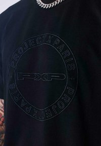 Camiseta negra con logo circular en relieve que dice "PROJECT X PARIS" alrededor de "PXP" en el centro, llevada con un collar de cadena plateada.