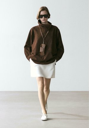 Person trägt eine übergroße braune Jacke, weißen Rock, weiße Schuhe, Sonnenbrille und eine braune Tasche, die am Hals hängt, und geht auf einem einfarbigen Hintergrund nach vorne.