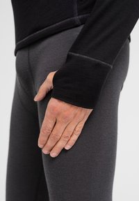 Sort langærmet bluse med kontraststing, parret med mørkegrå leggings. Hånden hviler på låret og viser stoffets struktur og pasform.