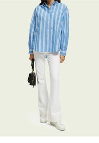 Blue and white striped button-up shirt with long sleeves, paired with white flared pants and white sneakers, holding a black handbag.