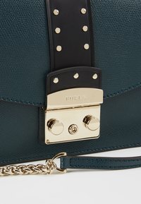 Borsa in pelle verde scuro con texture, tracolla in pelle nera, borchie dorate e una chiusura dorata incisa con "FURLA" e un dettaglio in catena dorata.