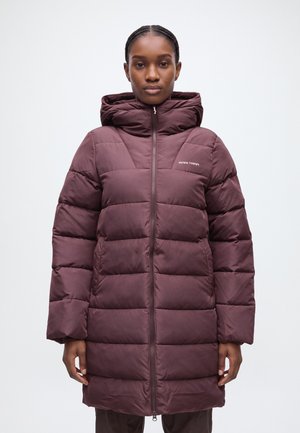 Une femme se tient face à l’objectif, vêtue d’un long manteau matelassé couleur prune avec une capuche haute et une fermeture éclair sur le devant, sur un fond clair et uni.