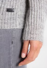 Pull gris en maille avec texture côtelée et une étiquette noire portant l'inscription "REPLAY" ; assorti à un pantalon gris, une main reposant sur le tissu.