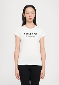Baltā īsaja piedurkne t-kreklā ir melns uzraksts "ARMANI EXCHANGE", kas centrēts uz krūtīm. Fit ir parasts, ar apaļu kakla izgriezumu.