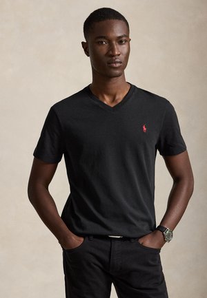 Ung mann iført svart V-hals T-skjorte med rødt logo og sorte bukser, stående med hendene i lommene mot ensfarget bakgrunn.