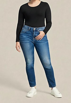 Personne portant un haut noir à manches longues, un jean bleu taille haute et des baskets blanches, debout avec une main dans la poche.