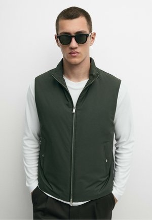 Jeune homme portant des lunettes de soleil noires, un gilet sans manches vert foncé zippé sur une chemise blanche à manches longues, debout devant un fond blanc.