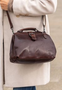 Sac à main en cuir marron au design structuré, avec une courte poignée et une bandoulière amovible. Il possède un fermoir en métal et des coutures visibles.