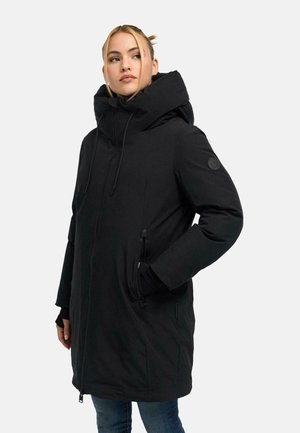 Femme portant un long manteau matelassé noir avec un col haut et une capuche, les mains dans les poches zippées, regardant légèrement sur le côté.