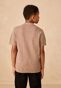 Tricou polo cu mâneci scurte, de culoare bej, cu un model geometric texturat, prevăzut cu un guler clasic. Purtați cu pantaloni negri.