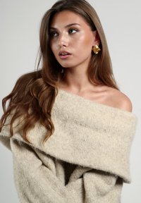 Suéter beige de gran tamaño en tejido suave con un amplio cuello off-shoulder. Los pendientes cuadrados dorados añaden contraste. Textura suave con tejido caído.