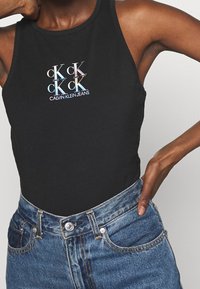 Musta hihaton bodysuit, jossa on keskellä kiiltävä monivärinen "CK"-logo. Yhdistetty sinisiin, korkeavyötäröisiin farkkuihin. Pehmeä, istuva materiaali.