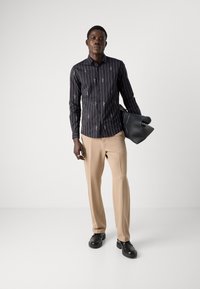 Camicia nera a maniche lunghe con strisce verticali bianche e motivi di foglie, abbinata a pantaloni beige e scarpe slip-on nere. L'uomo tiene una borsa nera.