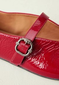 Red patent leather flat shoe with a smooth texture, featuring a single strap and silver buckle detail. Interior is tan leather.