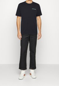Camiseta negra de manga corta con un sutil logo en turquesa, combinada con pantalones negros y zapatillas altas blancas. Los pantalones están doblados en los puños.