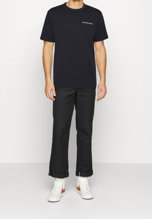Schwarzes T-Shirt mit kurzen Ärmeln und einem dezenten türkisfarbenen Logo, kombiniert mit schwarzen Hosen und weißen High-Top-Sneakern. Die Hosen sind an den Bündchen hochgekrempelt.