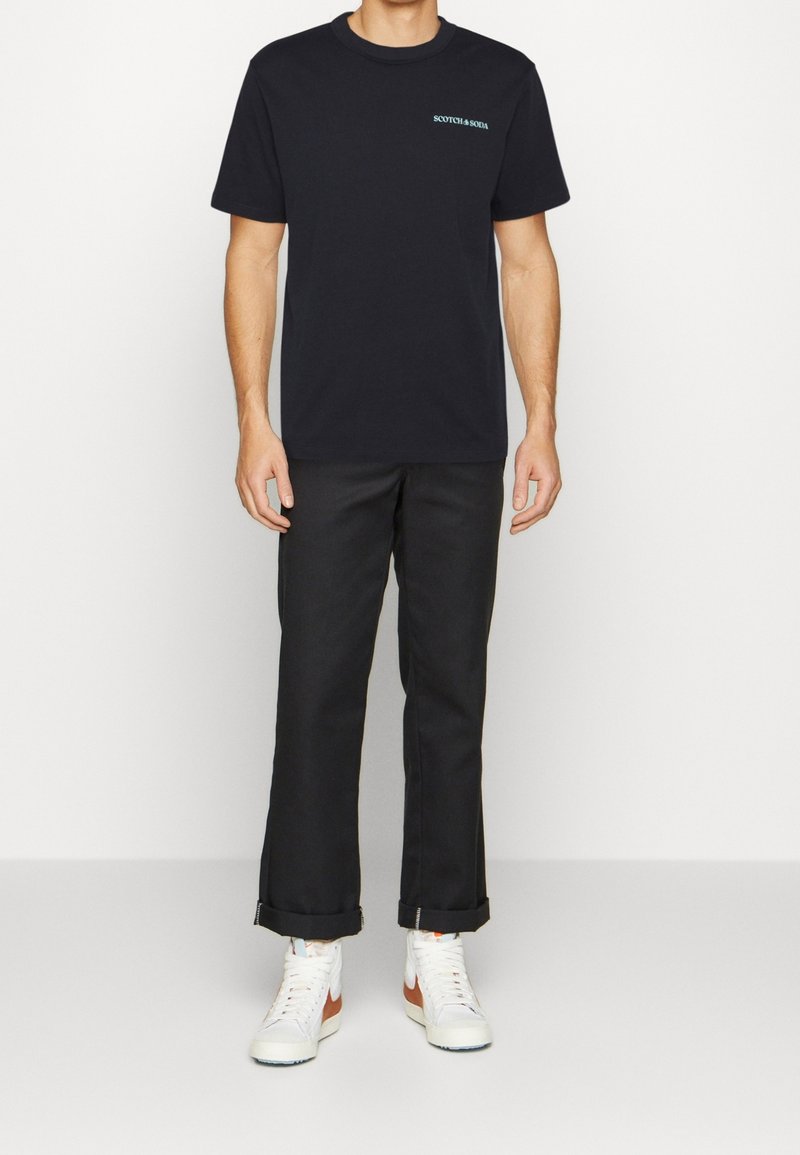 Camiseta negra de manga corta con un sutil logo en turquesa, combinada con pantalones negros y zapatillas altas blancas. Los pantalones están doblados en los puños.