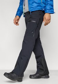 Ανδρικό μαύρο παντελόνι υπαίθριου τύπου με τσέπες με φερμουάρ, ενισχυμένα κάτω μέρη και στενή εφαρμογή, σε συνδυασμό με μαύρα πεζοπορικά παπούτσια.