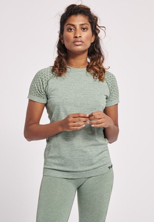 Frau mit lockigem Haar, die ein enganliegendes, hellgrünes Sportshirt mit kurzen Ärmeln und passende Leggings trägt, steht vor einem schlichten Hintergrund.