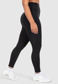 Leggings neri a vita alta con una texture liscia e elastica. Indossati con scarpe da ginnastica bianche con suola spessa e dettagli neri.