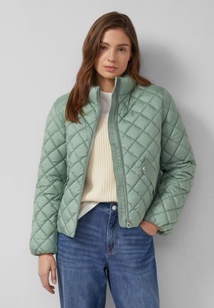 Noor naine, kes kannab helerohelist tikitud jopet, mis on osaliselt tõmmatud üle kreemika värvi kampsuni ja siniste teksapükste, seisab halli tausta ees.