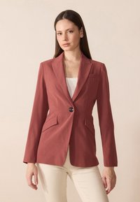Rustrød blazer laget av glatt stoff med slim fit, notched revers, en enkelt frontknapp, og to gentekkerlommer. Enfarget bakgrunn.