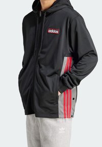 Giacca nera con cappuccio con dettagli rossi e grigi, chiusura con zip, tasche anteriori e logo Adidas. Abbinata a pantaloni della tuta grigi.