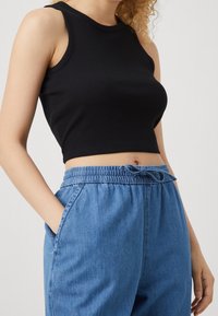 Woman wearing a sleeveless black crop top and high-waisted blue denim jogger pants with a drawstring, hands in pockets.