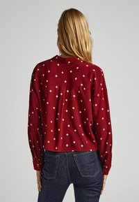 Camisa roja de manga larga con un sutil estampado de diamantes en color crema. Está hecha de una tela suave, tiene un corte recto y puños abotonados.