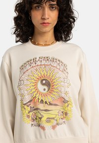 Krämfärgad sweatshirt med en flerfärgad grafik av en sol, yin-yang symbol och blommor. Rund hals och avslappnad passform.