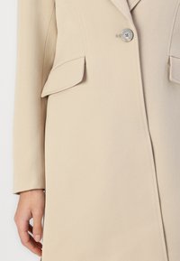 Manteau beige avec une fermeture à bouton unique, deux poches à rabat et des manches longues au design minimaliste et à la texture lisse.