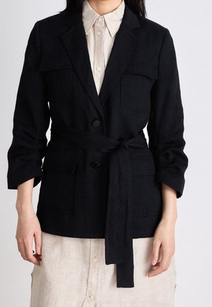Kvinde iført en sort blazer med bælte og klaplommer over en beige kjole med knapper, ærmerne rullede op til underarmen.