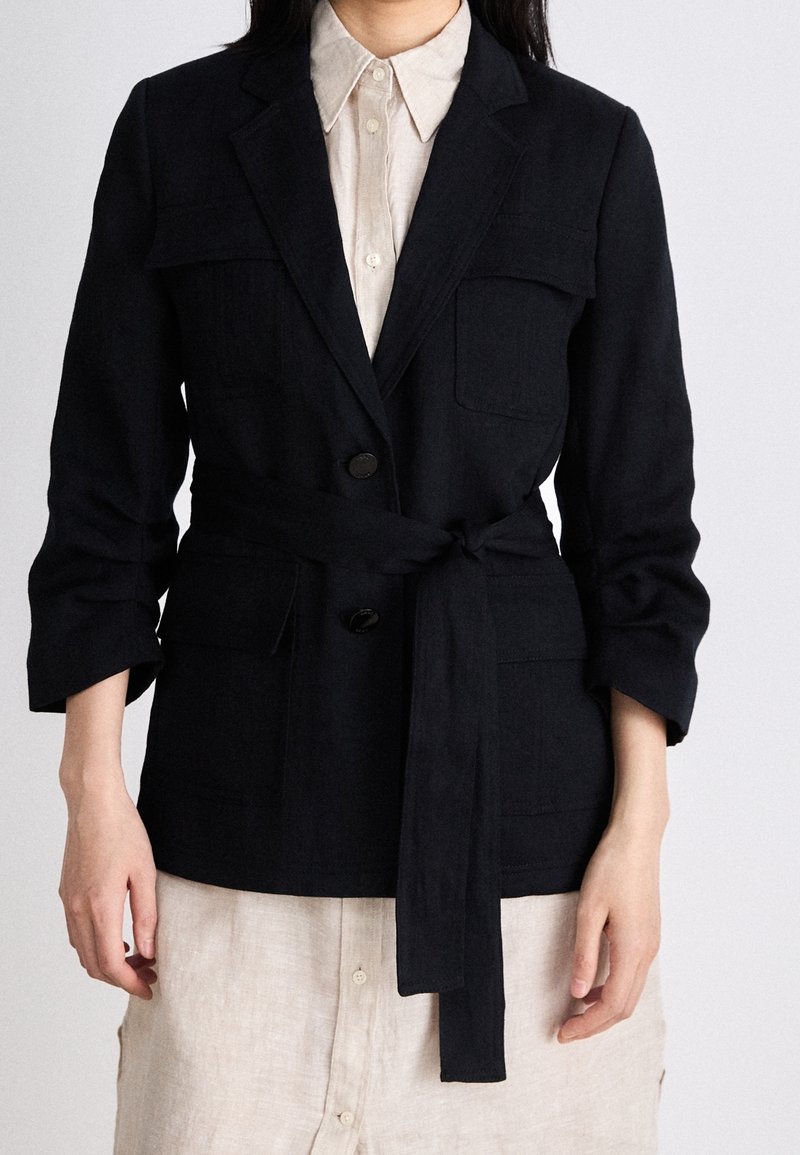Femme portant un blazer noir ceinturé avec poches à rabat sur une robe beige boutonnée, manches retroussées jusqu'à l'avant-bras.