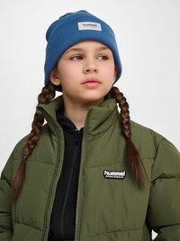 Kind mit braun geflochtenem Haar trägt eine blaue Mütze und eine grüne Daunenjacke mit schwarzem Reißverschluss und hummel Sportlogo.