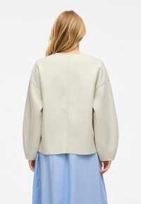 Šviesiai smėlinis megztinis su per didelėmis, nuleistomis rankovėmis ir laiveliu iškirpte. Minkšta tekstūra, švarios linijos ir matomi siūlių detalizavimai.